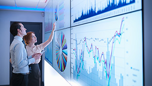 Two business professionals analyzing financial charts and graphs displayed on large monitors.