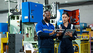 Two manufacturing workers in a factory reviewing information on a digital tablet beside industrial machinery.